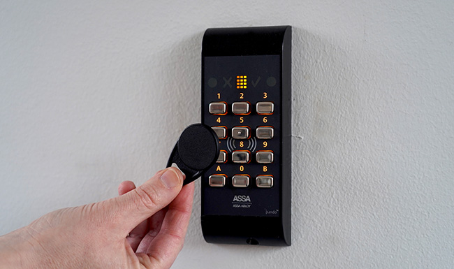 Svart kortläsare, dosa med knappar, på vit vägg samt en hand som sträcker fram en svart nyckeltagg.