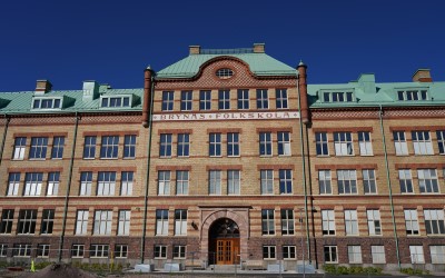 Skolbyggnad i tegel, många fönster och entré samt texten Brynäs Folkskola står på fasaden.