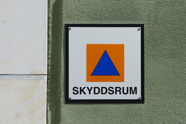 Skylt med orange kvadrat och blå triangel på vit botten, med texten Skyddsrum. Sitter på grön bakgrund.